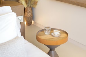 A neatly made bed with white linens, a small round wooden table with a glass of water and a cup, and a wall-mounted control panel. - Avgoustos Suites - Adults only (Naxos)