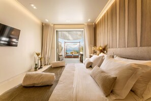A modern hotel room with a large bed, a TV mounted on the wall, and a view of the outdoors through a glass door. - Avgoustos Suites - Adults only (Naxos)