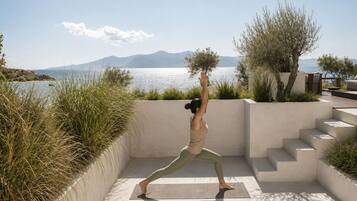 Una persona realiza una postura de yoga sobre una esterilla, con vistas a un cuerpo de agua y montañas al fondo.
