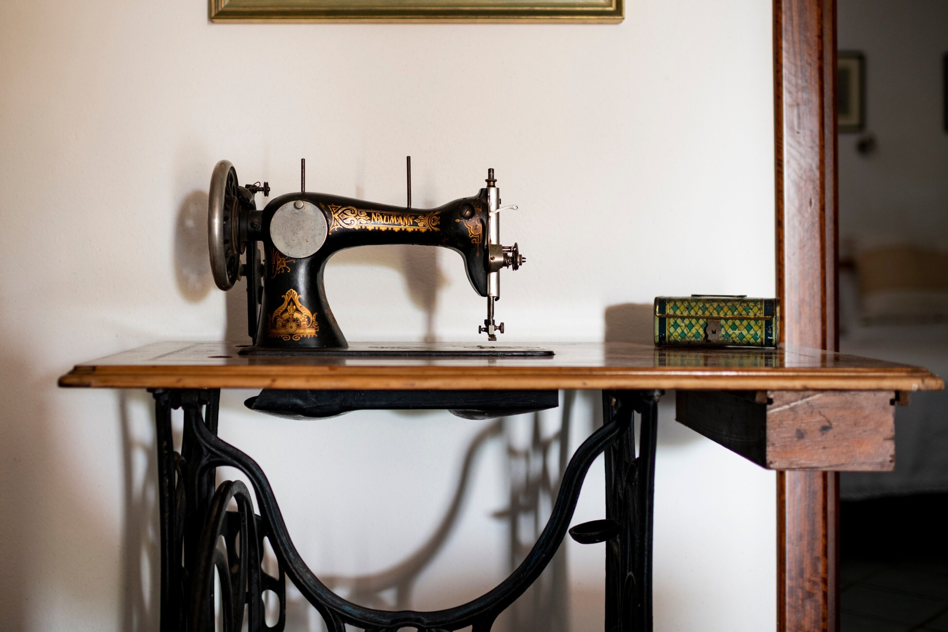 Una máquina de coser sobre una mesa con tapa de madera y estructura de metal negro, una caja verde con diseño estampado y un cuadro en la pared.