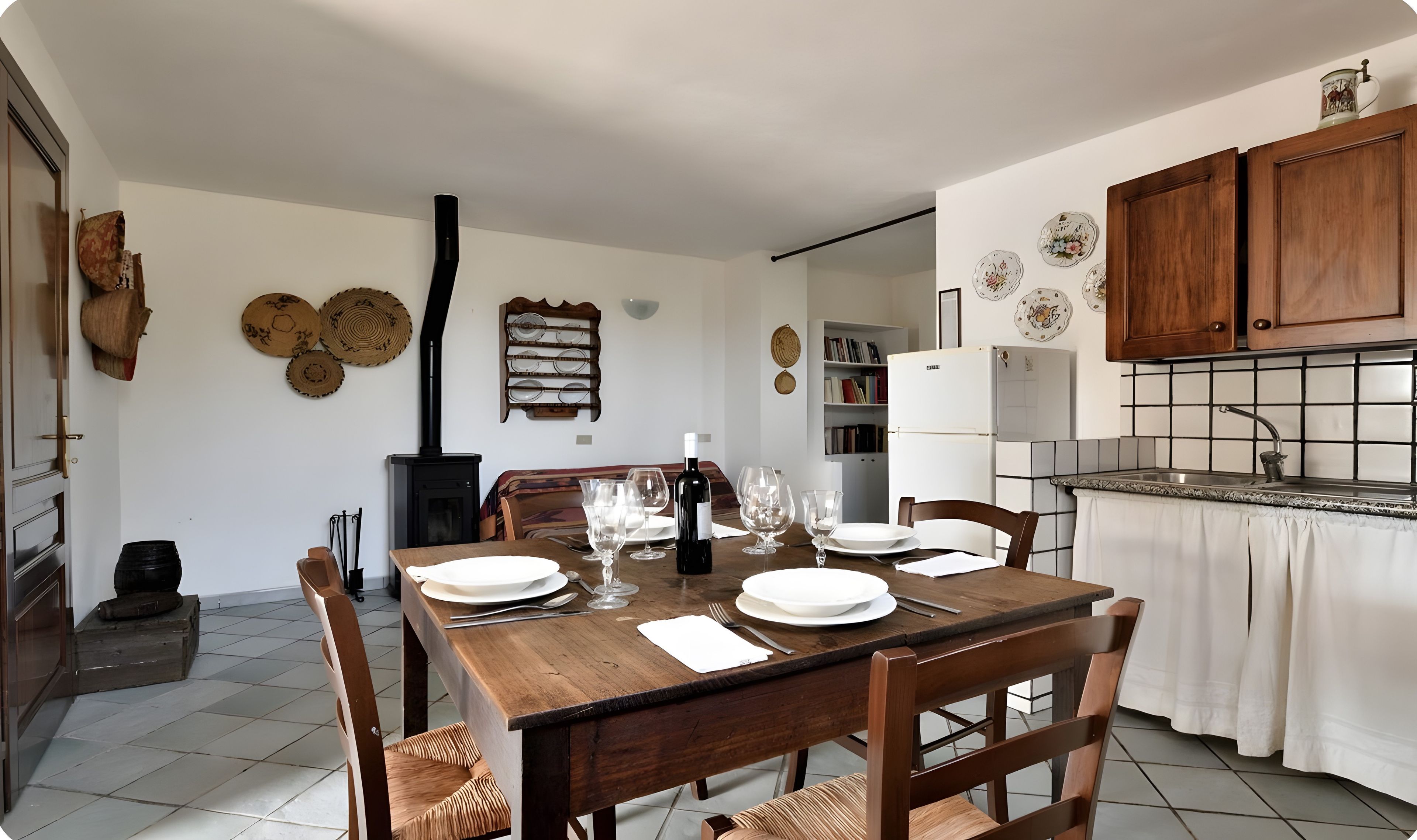 Un coin repas avec une table en bois dressée pour quatre personnes, un poêle noir et une cuisine dotée de meubles en bois et d’un dosseret carrelé.