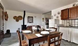 Un coin repas avec une table en bois dressée pour quatre personnes, un poêle noir et une cuisine dotée de meubles en bois et d’un dosseret carrelé.