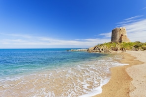 A coastal scene with a stone tower on a rocky outcrop, clear blue water, and a sandy beach.