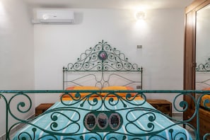 A bedroom with a metal bed frame, a single bed with an orange and blue bedspread, a small wooden nightstand, a wall-mounted air conditioner, and a wooden wardrobe.