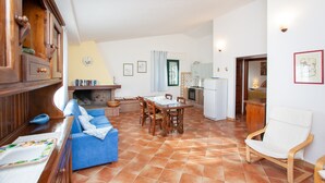 A spacious living area with a blue sofa, wooden dining table, and chairs, a kitchen in the background, and a white chair near the entrance. - Holiday Home Casa Barvis with Garden, A/C, Terrace and Wi-Fi (Orosei)