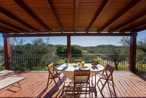 A covered outdoor dining area with a table set for two, surrounded by chairs, overlooking a scenic landscape.