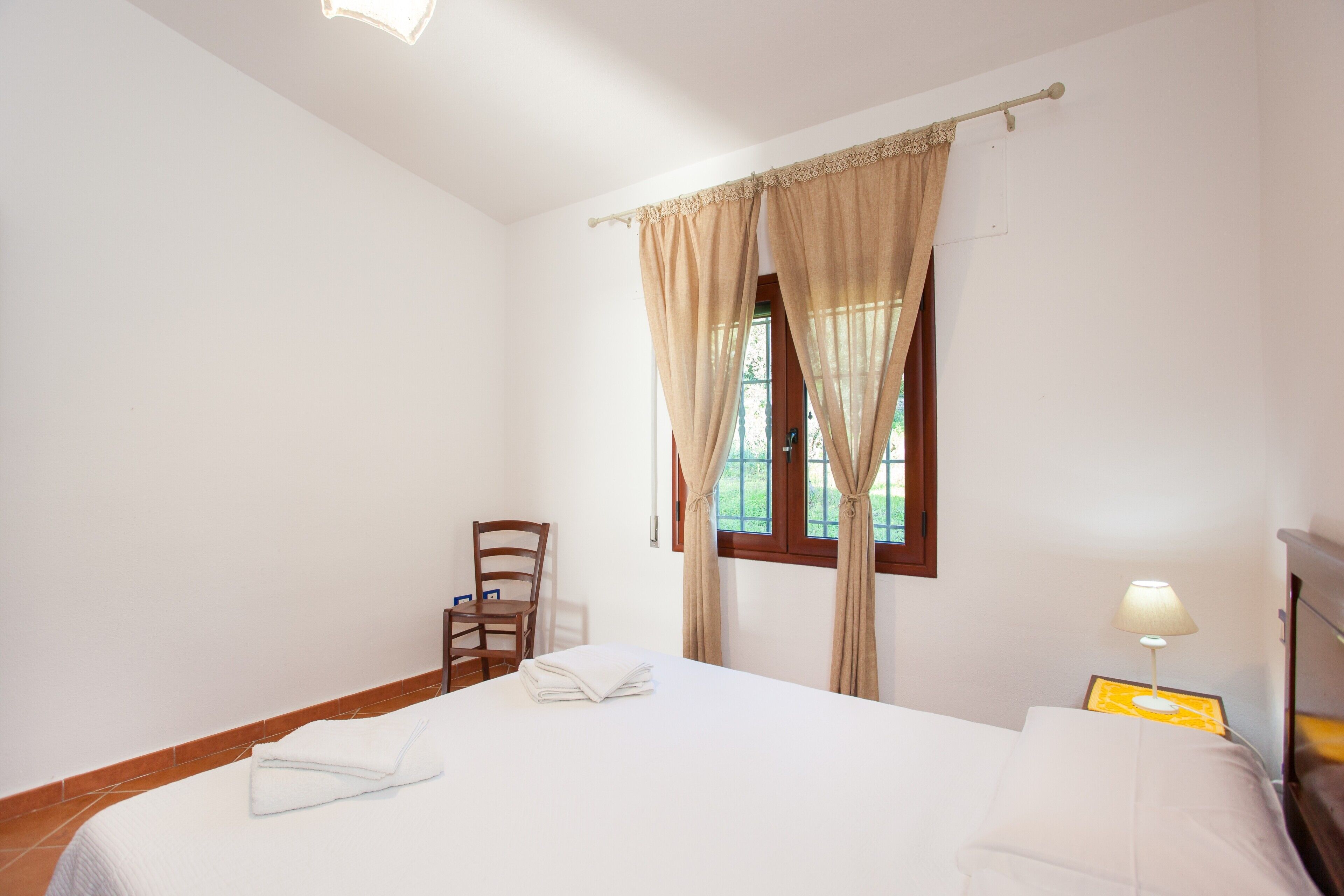 Une chambre d’hôtel avec un lit, une chaise, une fenêtre avec des rideaux, une table de chevet avec une lampe et une porte.