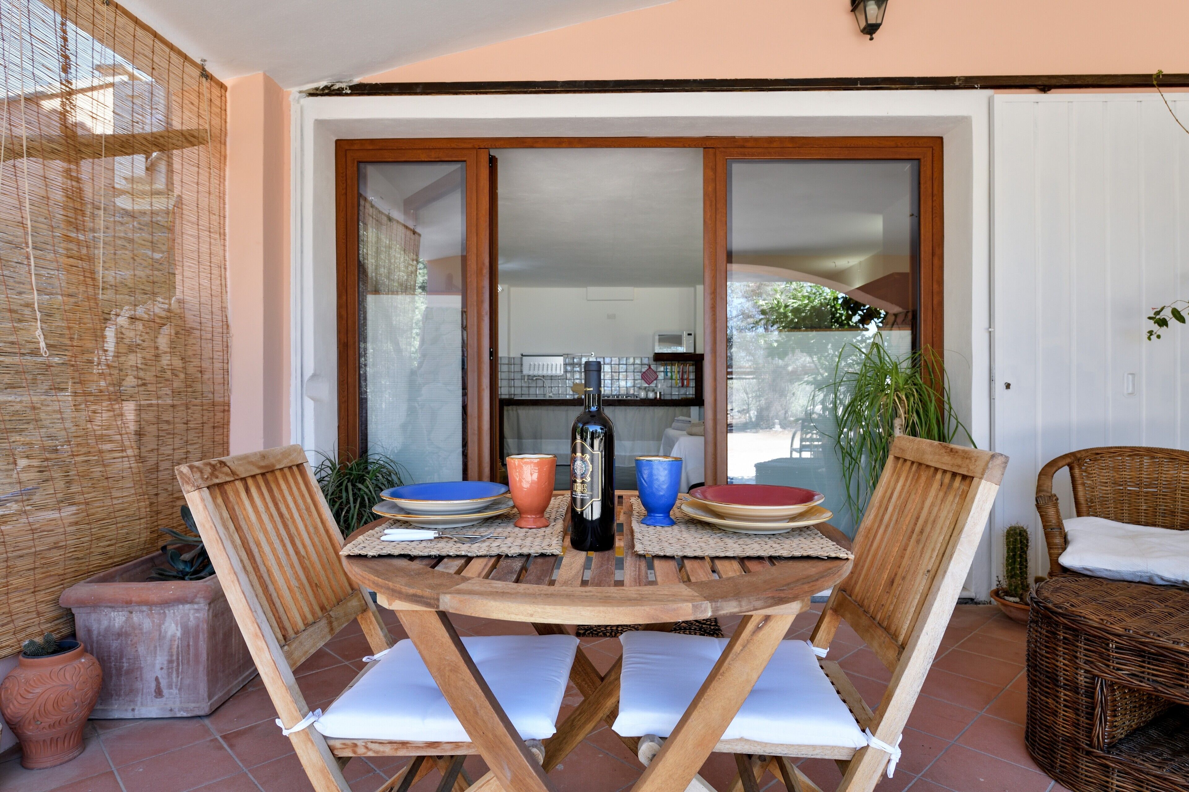 A wooden table with chairs, a bottle, and plates on a patio with a view of the interior.