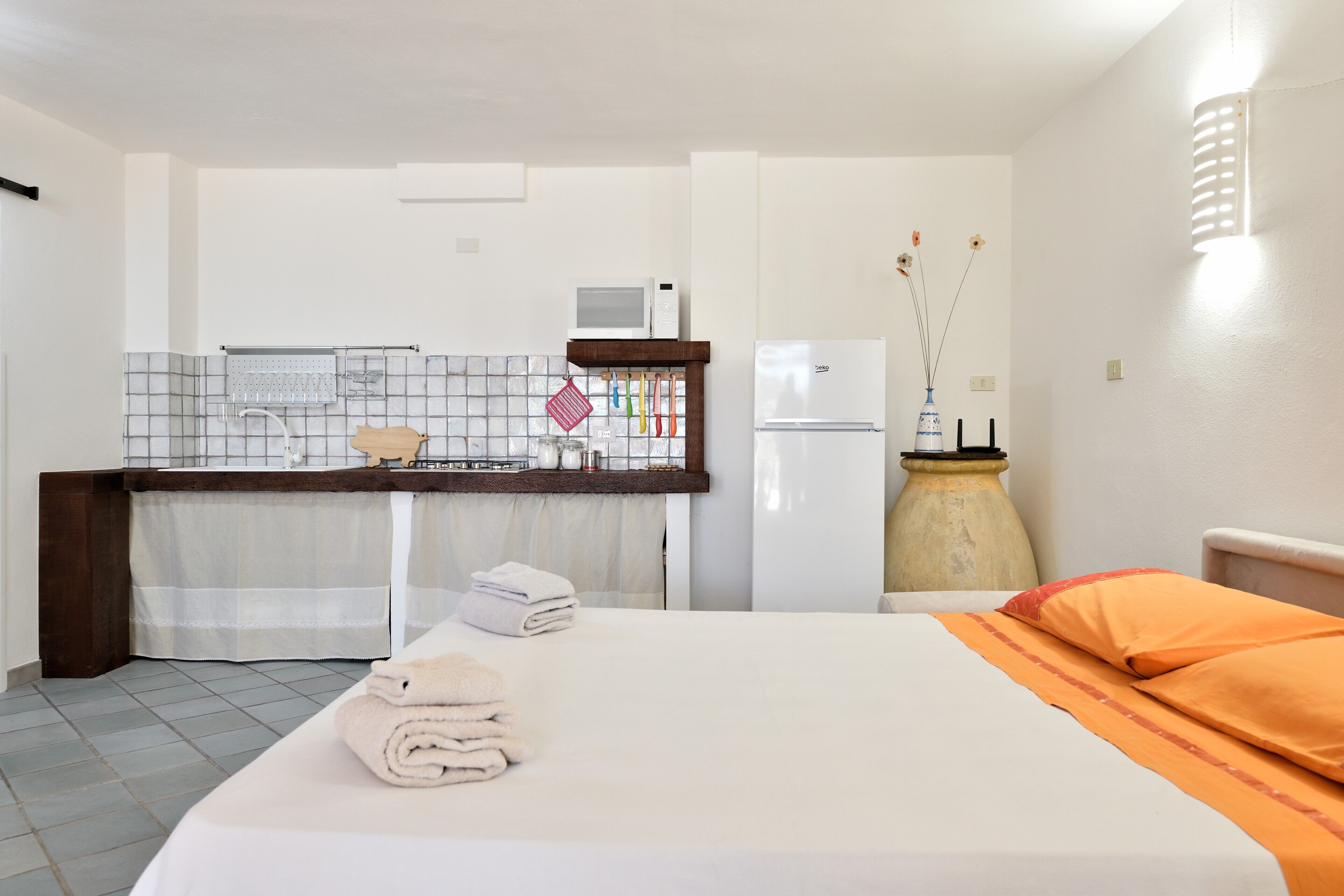 Hotellihuone, jossa on sänky, pieni keittokomero, mikroaaltouuni, jääkaappi ja maljakko, jossa on kukkia.