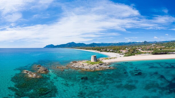 Paisagem costeira com águas turquesa cristalinas, uma praia arenosa e um promontório rochoso com uma torre.