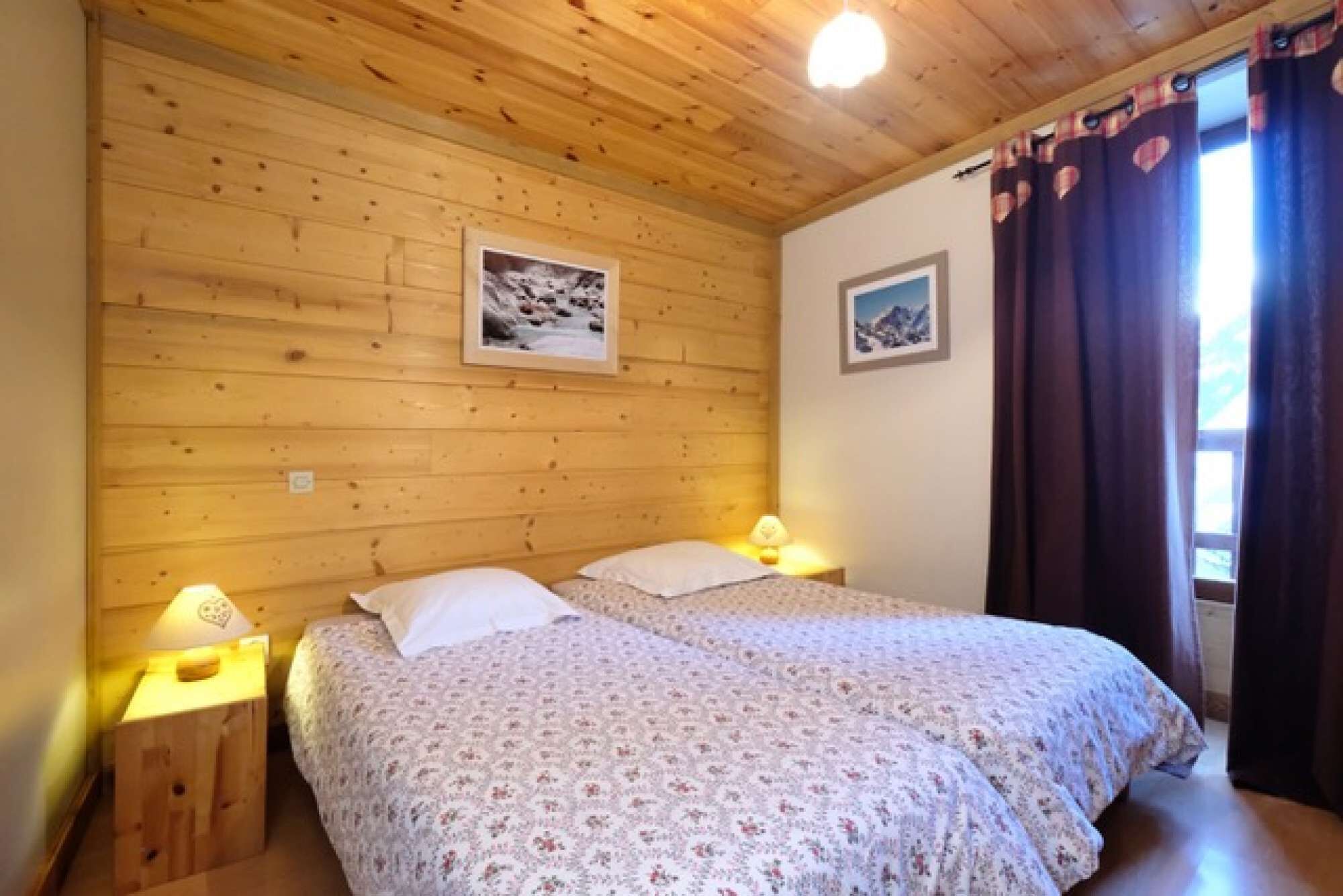 Quarto com paredes de madeira, duas camas com roupa de cama florida, duas luminárias de cabeceira e duas imagens emolduradas.
