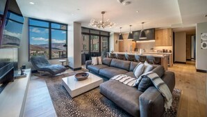 A modern living room with a large sectional sofa, a flat-screen TV, and a dining area with a kitchen in the background. - One Snowmass East 303 (Snowmass)
