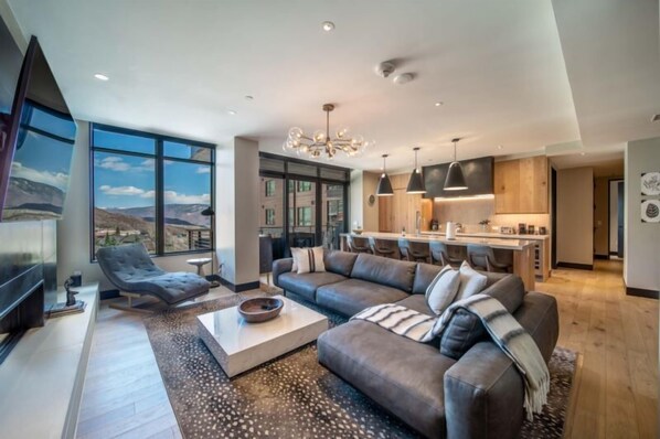 A modern living room with a large sectional sofa, a flat-screen TV, and a dining area with a kitchen in the background. - One Snowmass East 303 (Snowmass)