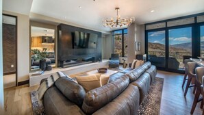 A modern living room with a large sectional sofa, a flat-screen TV, and a dining area with a view of the desert landscape. - One Snowmass East 303 (Snowmass)