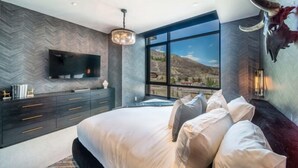 A modern bedroom with a large bed, a TV mounted on the wall, and a view of the mountains through large windows. - One Snowmass East 303 (Snowmass)