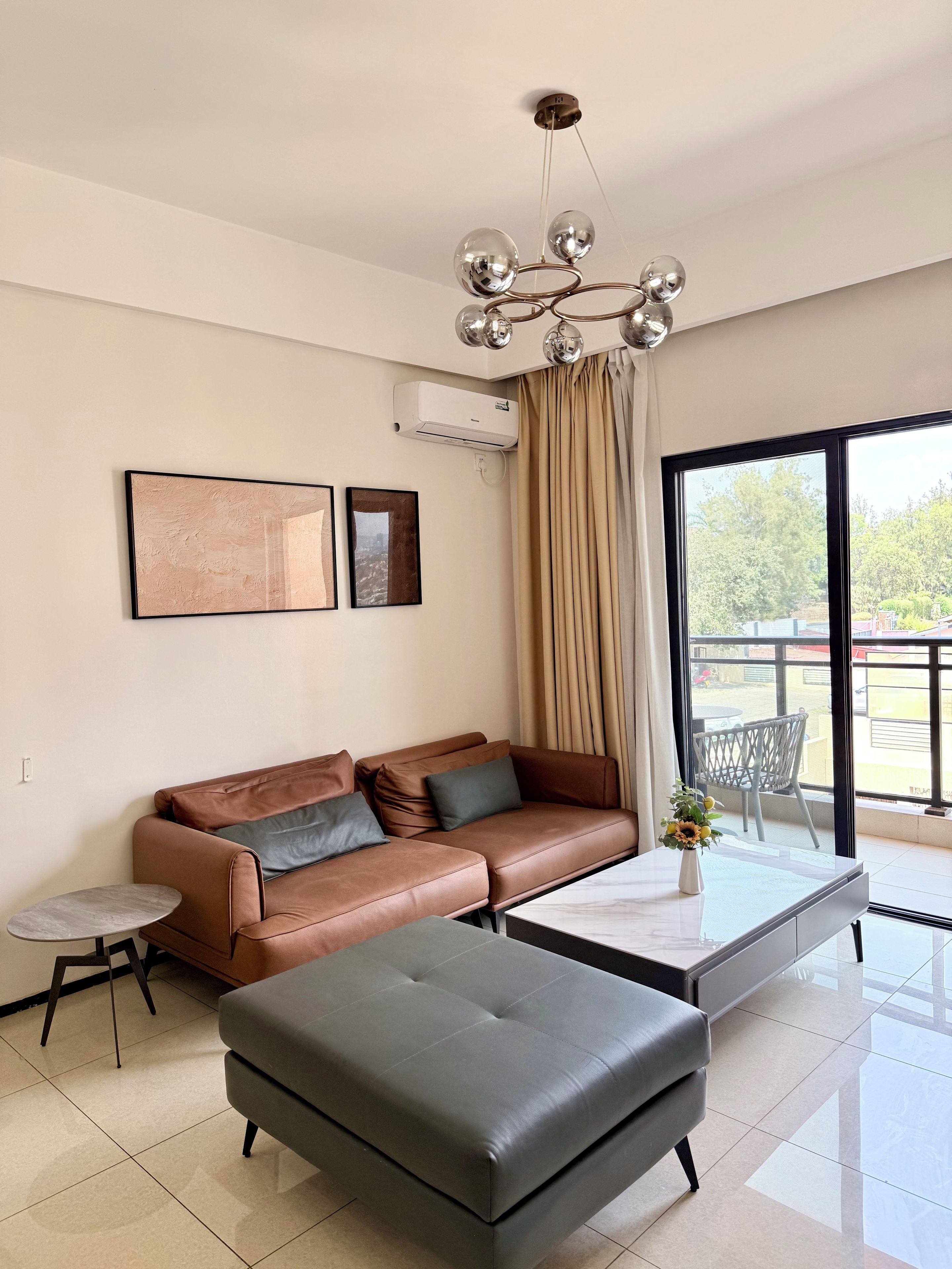 Ein modernes Wohnzimmer mit einem Ledersofa, einem grauen Hocker, einem kleinen Beistelltisch und einem großen Fenster mit Blick ins Freie.