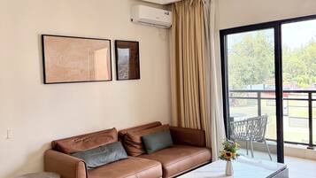 Ein modernes Wohnzimmer mit einem Ledersofa, einem grauen Hocker, einem kleinen Beistelltisch und einem großen Fenster mit Blick ins Freie.