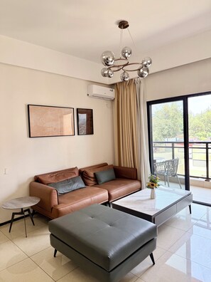 Een moderne woonkamer met een leren bank, een grijze poef, een klein bijzettafeltje en een groot raam met uitzicht op de omgeving.