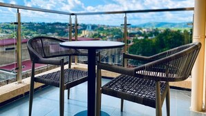 Uma varanda com uma mesa redonda e duas cadeiras de vime, com vista para uma paisagem urbana com edifícios e áreas verdes.