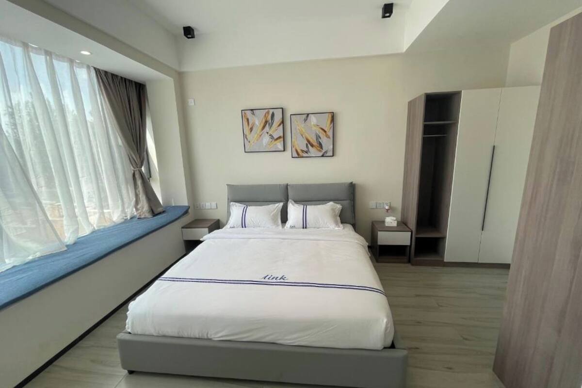 A modern hotel room with a large bed, a bedside table, a wardrobe, and a window with sheer curtains.