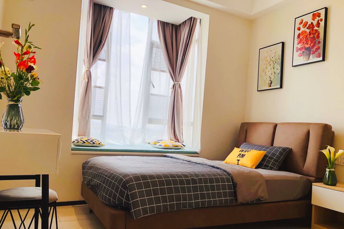 A bedroom with a bed, a window with curtains, a bedside table, a chair, and a vase with flowers.