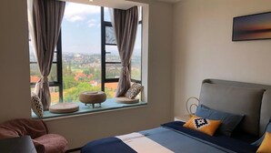 Quarto com uma janela grande, cama com roupa de cama azul e branca, e vista para um horizonte urbano.
