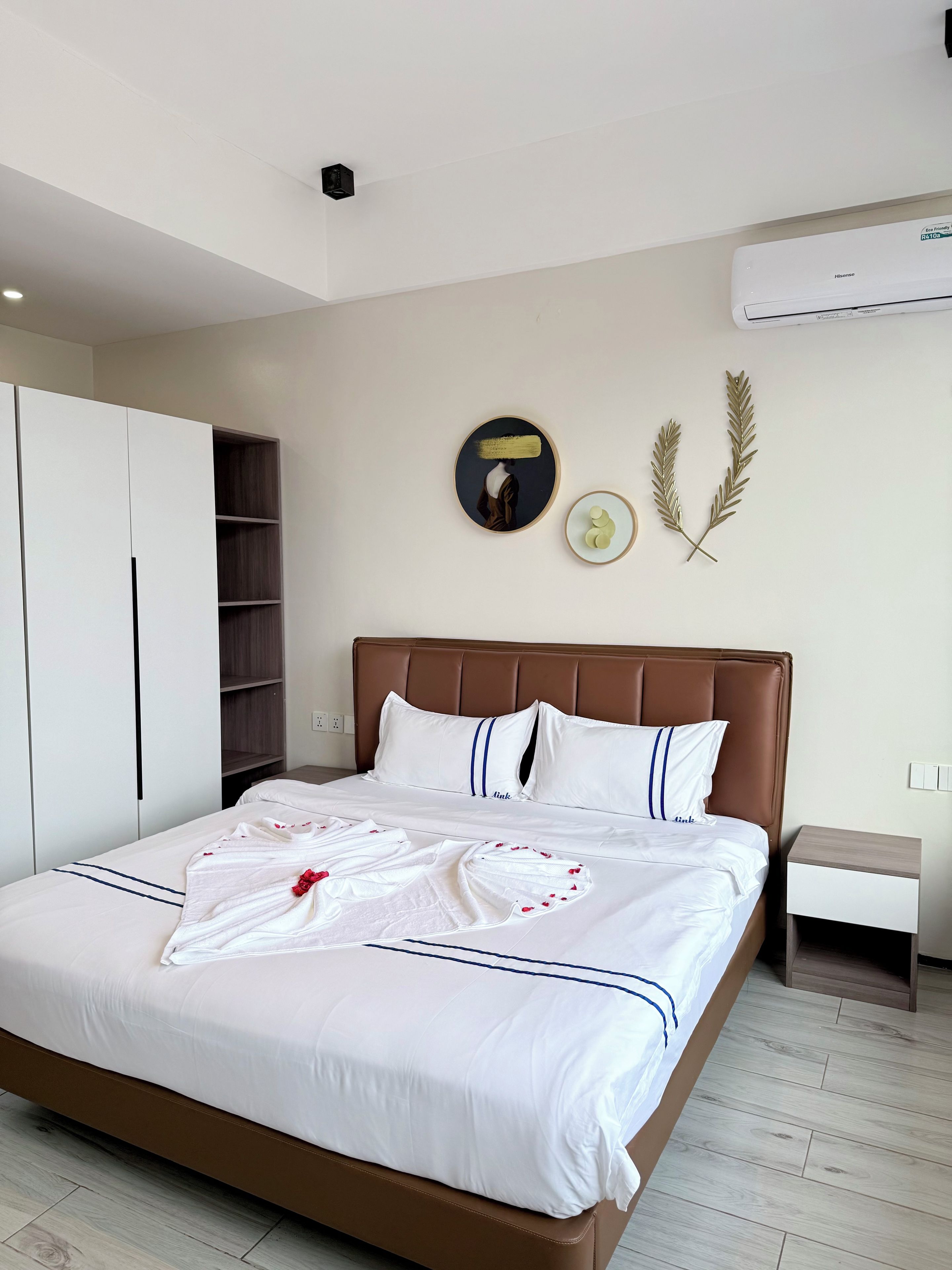A neatly made bed with white bedding and decorative pillows, a brown headboard, and a bedside table with a lamp.