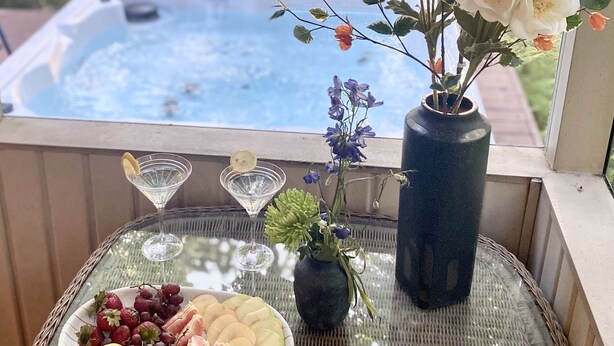 Uma mesa de vidro com uma travessa de frutas, um vaso com flores e uma banheira de hidromassagem ao fundo.