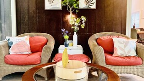 Um espaço de estar aconchegante com cadeiras de vime e almofadas vermelhas, uma pequena mesa lateral com um vaso de flores e obras de arte emolduradas na parede.
