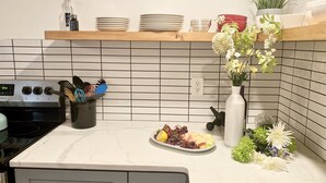 Uma cozinha com fogão, pia e bancada com um prato de frutas. Há pratos, um vaso com flores e uma planta em vaso na bancada.