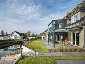 Ein modernes Haus mit Wintergarten, einem gepflasterten Weg und einem kleinen Anlegesteg an einem Kanal.