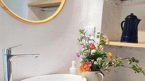 Ein Badezimmer mit rundem Spiegel, Waschbecken in Weiß, Holzwaschtisch, einer Topfpflanze und einem Regal mit Wasserkocher.