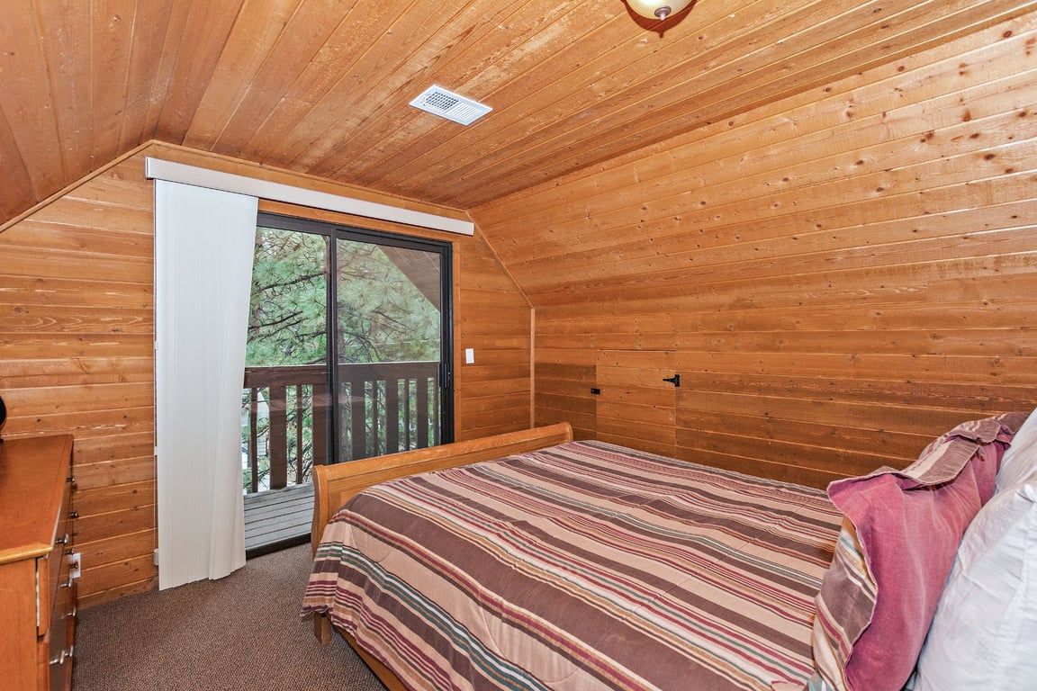 A bedroom with a wooden ceiling, a bed with a striped bedspread, a window with a view of trees, and a door leading to an outdoor deck.