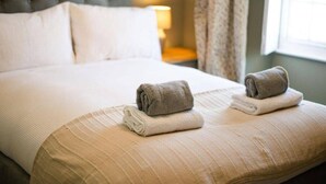 Un lit bien fait, avec deux serviettes pliées posées sur les oreillers, une tête de lit capitonnée, une lampe de chevet et une fenêtre avec des rideaux.