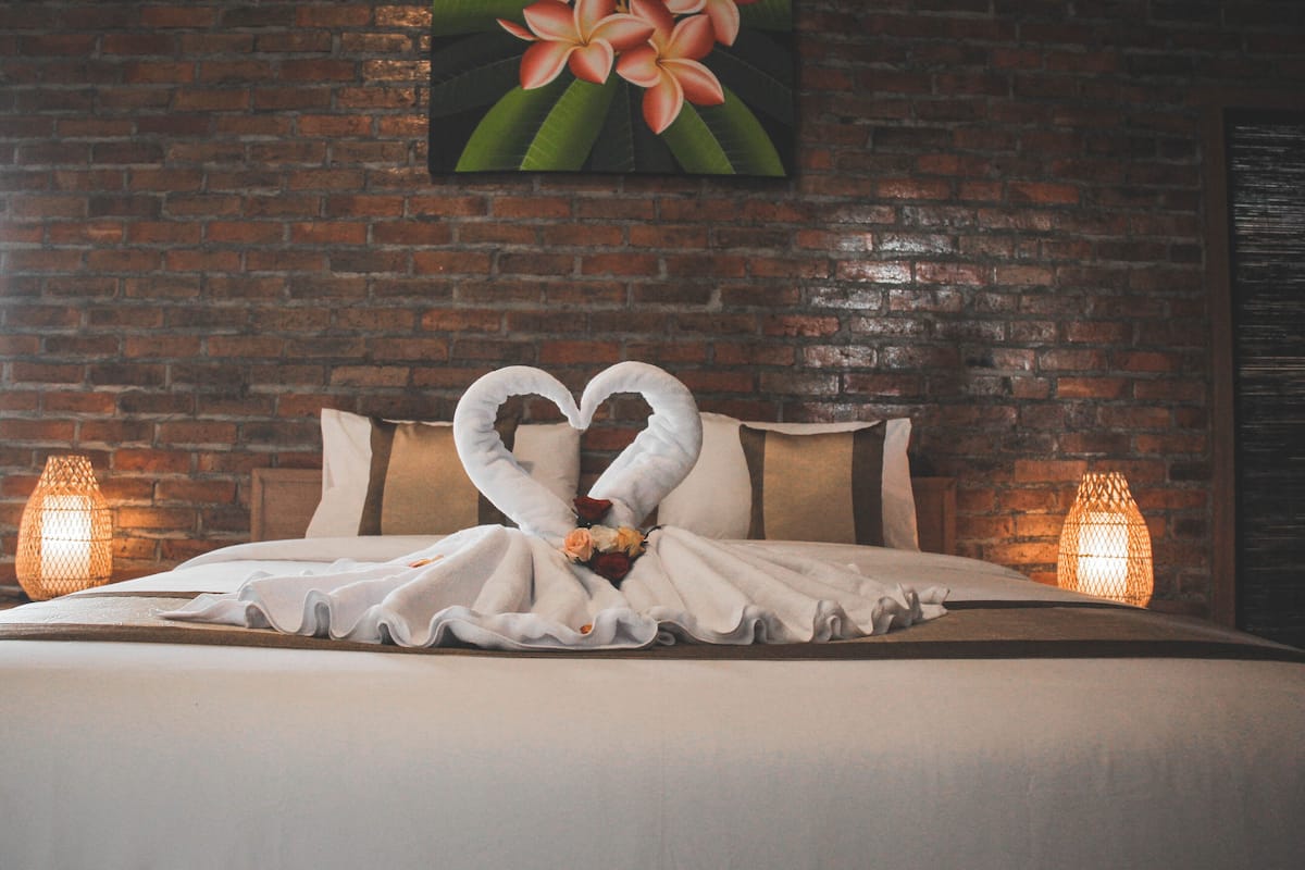 A hotel room with a brick wall, a bed with a heart-shaped towel arrangement, and two wall-mounted lamps.
