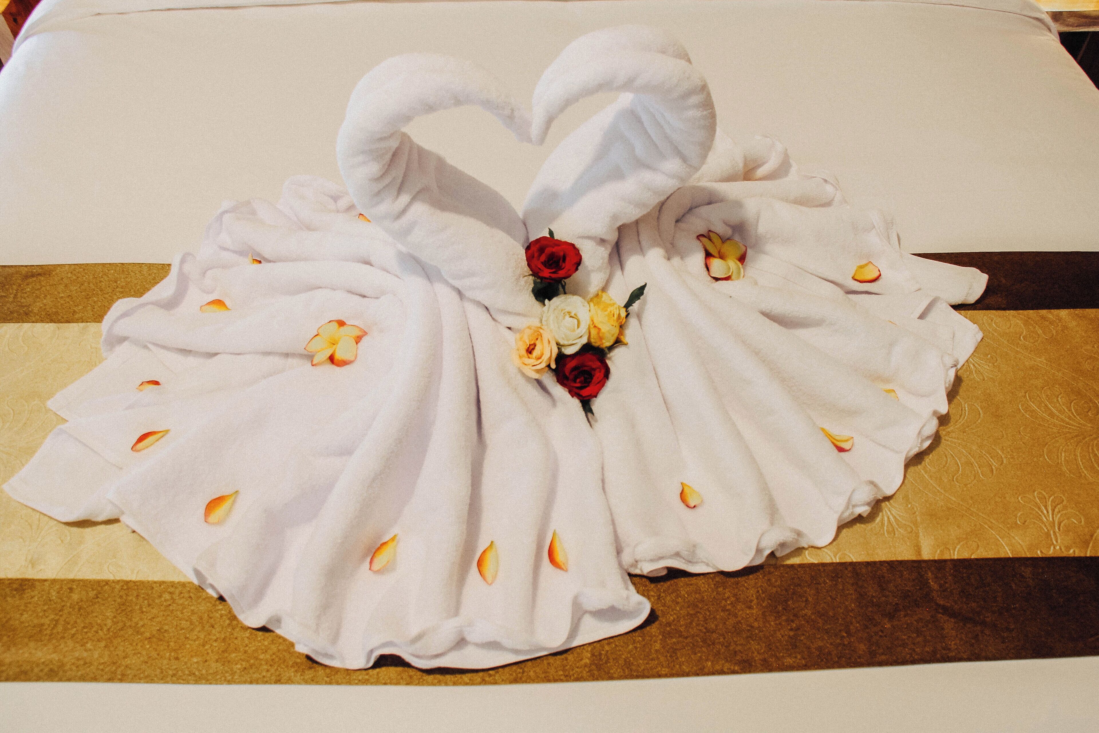 A bed with a white towel arranged in a heart shape, adorned with flowers and rose petals.