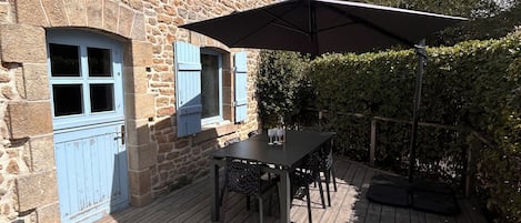Un bâtiment en pierre avec des volets bleus, une terrasse en bois et une table entourée de chaises.