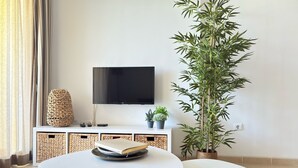 Un salon moderne avec un téléviseur à écran plat, une grande plante en bambou et une table ronde blanche accompagnée de deux chaises.