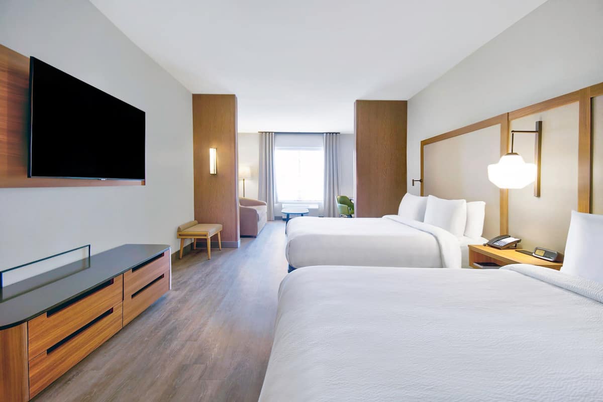 A hotel room with two beds, a flat-screen TV mounted on the wall, a desk with a chair, and a bedside table with a lamp.