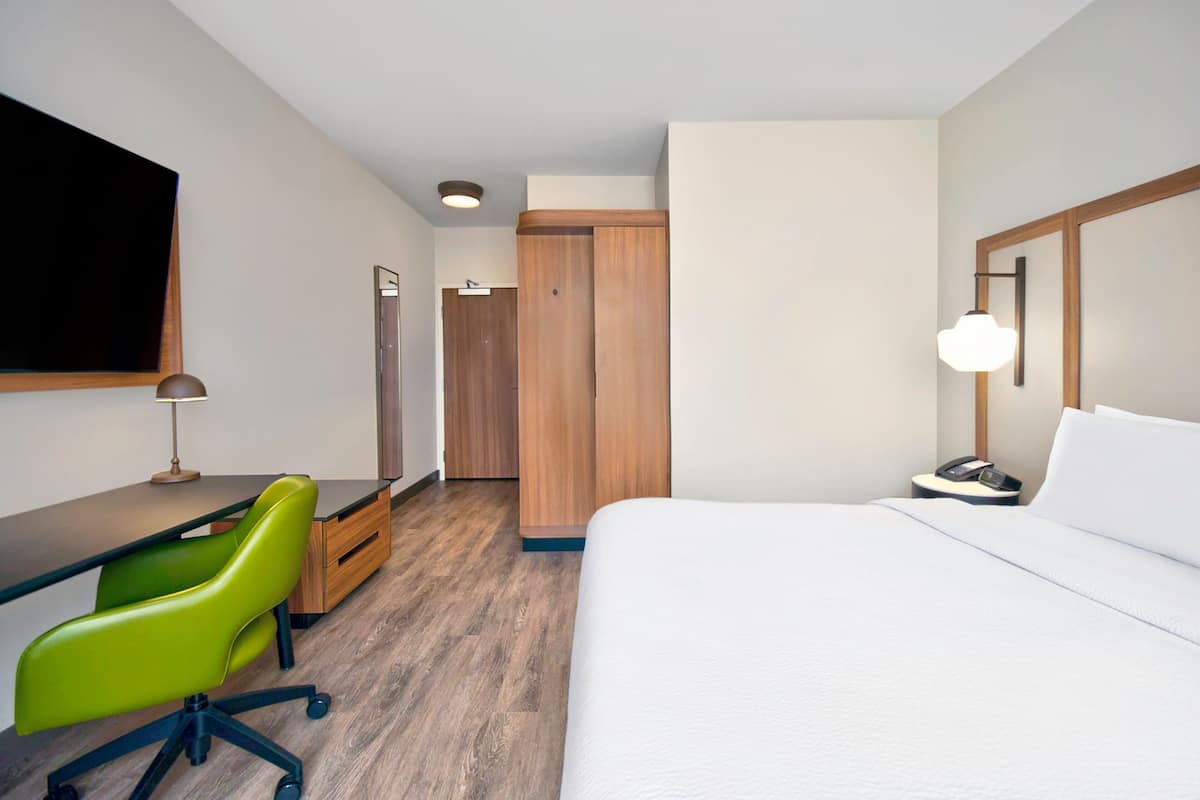 A hotel room with a bed, a desk with a chair, a wardrobe, and a bedside table with a lamp.