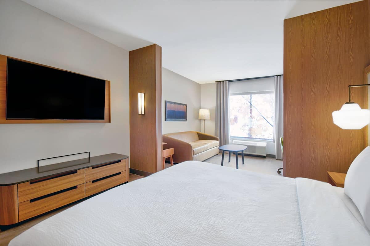 A hotel room with a large bed, a TV mounted on the wall, a sofa, a small table, and a window with curtains.