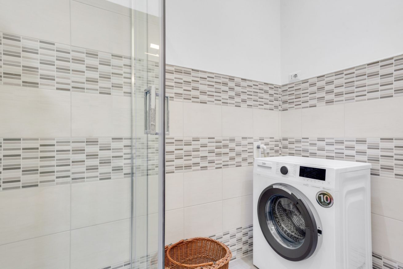 Un baño moderno con cabina de ducha de vidrio, lavadora blanca y cesta de mimbre.