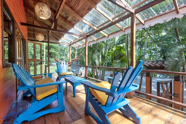 A covered outdoor seating area with blue chairs and yellow cushions, surrounded by glass panels and tropical plants. - Beachfront X T Sea - Surf Spot with AC and FiberOp (Cocles)