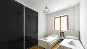 A modern bedroom with a sliding glass door, two beds, a bedside table, a hanging light fixture, and a window with blinds.
