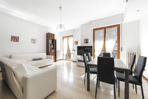 A modern living room with a white sofa, a dining table with chairs, a TV, and a bookshelf.