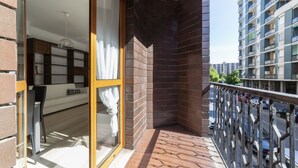 A balcony with a view of a cityscape, a glass door leading to a room with a sofa and a table, and a brick wall.