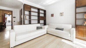 A modern living room with a white sofa, wooden shelves, and a dining area visible in the background.