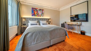 Ein Hotelzimmer mit einem großen Bett, einem Schreibtisch, einem Stuhl, einem Fernseher und einem farbenfrohen Gemälde an der Wand.