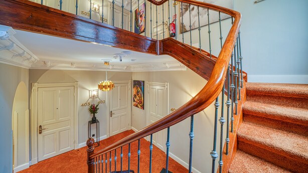 Eine imposante Treppe mit hölzernem Geländer und blauen Spindeln, die zu einer Zwischenebene mit einer Chandelieber und einer Tür mit der Aufschrift „403“ führt.