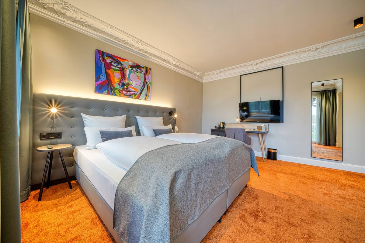 Una habitación de hotel moderna con una cama grande, una mesita de noche, un escritorio, un espejo y un cuadro llamativo en la pared.
